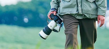 A person walking through a field while holding a camera with a super-telephoto lens