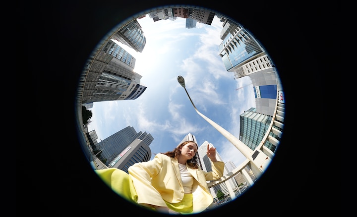 Circular fisheye view of a person surrounded by buildings, with the architecture appearing to lean inward toward the center of the frame