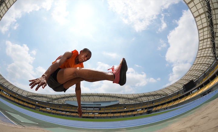 Diagonal fisheye view of a long jumper in motion, capturing the expansive stadium interior and ceiling within a single frame